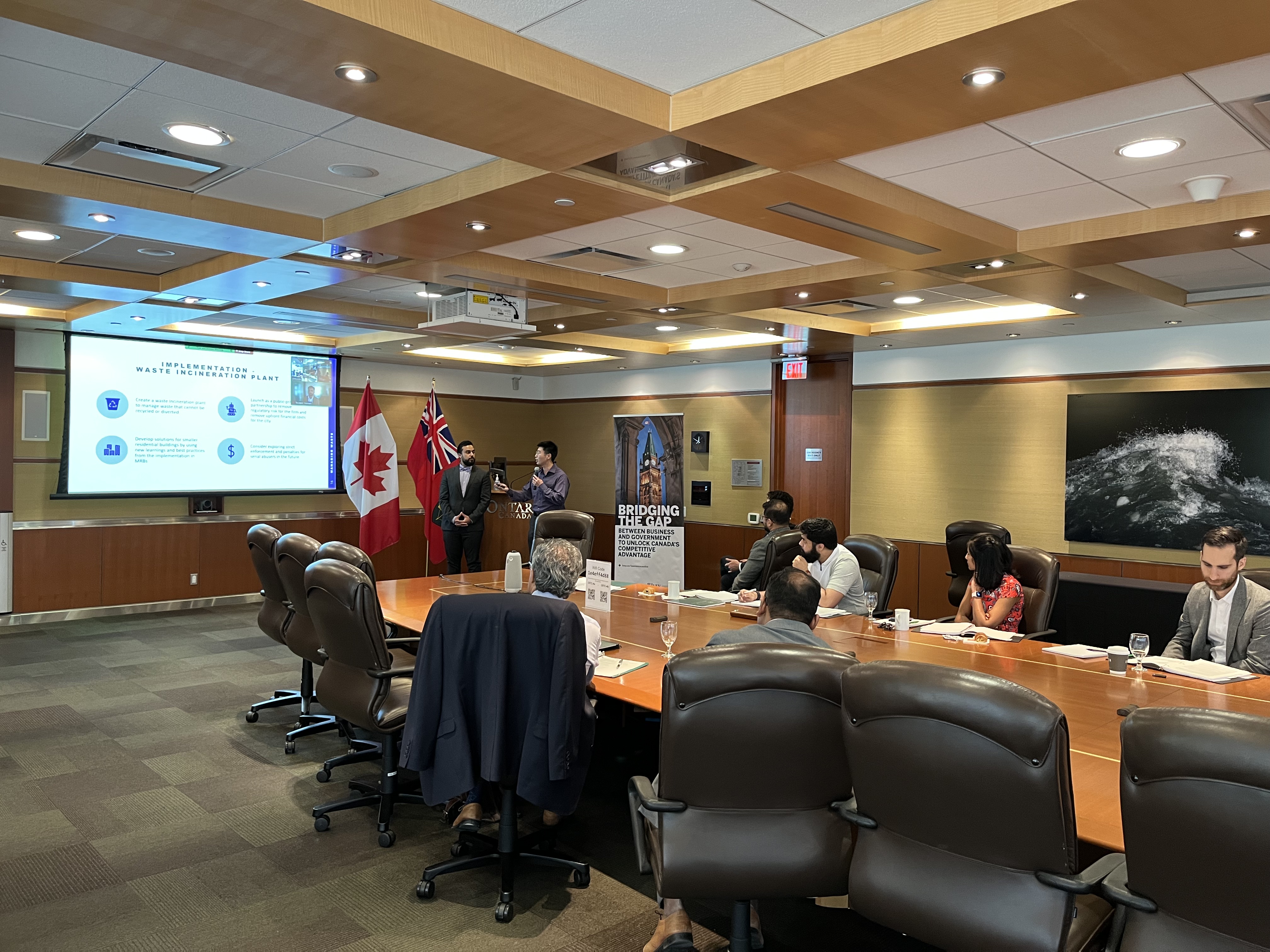 Two students stand at the front of a large boardroom, to the right of a drop down screen. Power point presentation is on the screen, along with a Zoom video of third student participating virtually