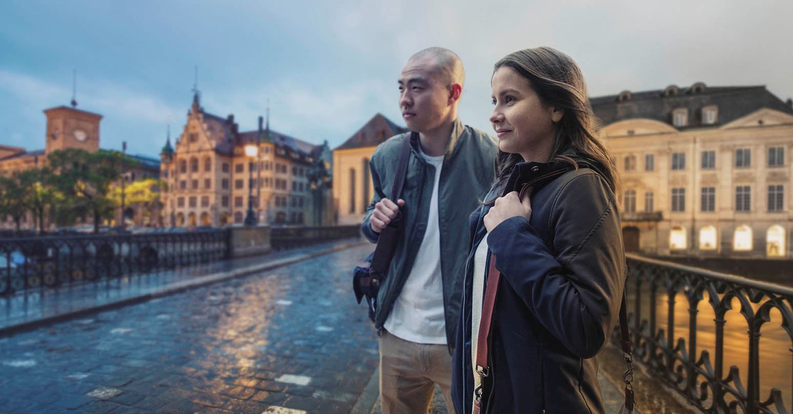 A young man and young woman walking together in Geneva with water and buildings in the background