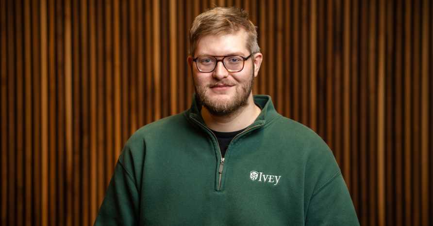Photo Of Ethan Milne Ivey Phd Candidate Wearing Green Ivey Branded Sweater In Wood Paneled Room