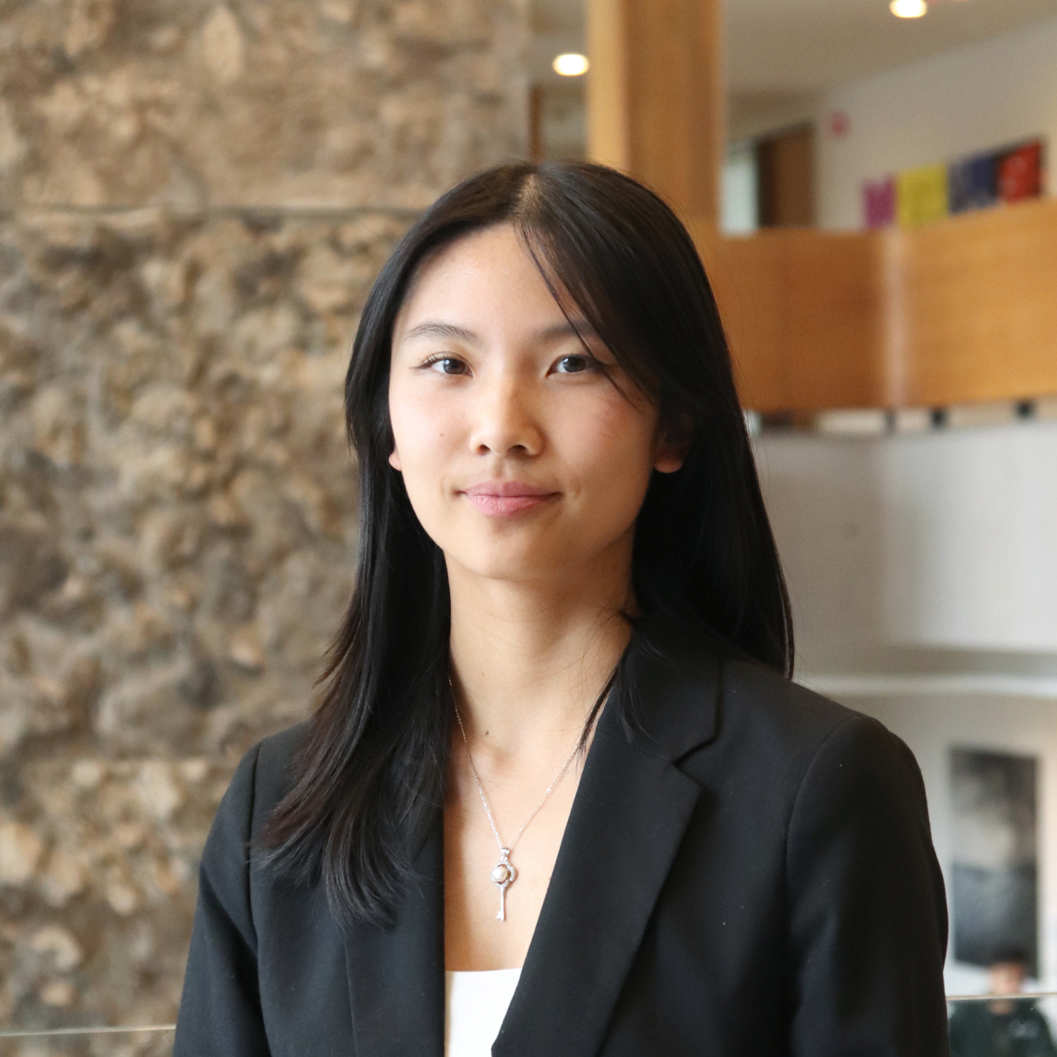 Headshot of Laurel Dong, an Ivey HBA student.  Laurel has shoulder length, straight black hair and wears a black blazer with a white tee shirt.  The main floor of Ivey is blurred in the background.