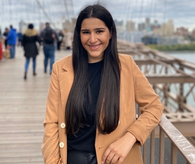 Headshot of MBA Student Ayesha Zaman. She is standing on a bridge, posing in a light brown jacket and black shirt. She has long black hair.