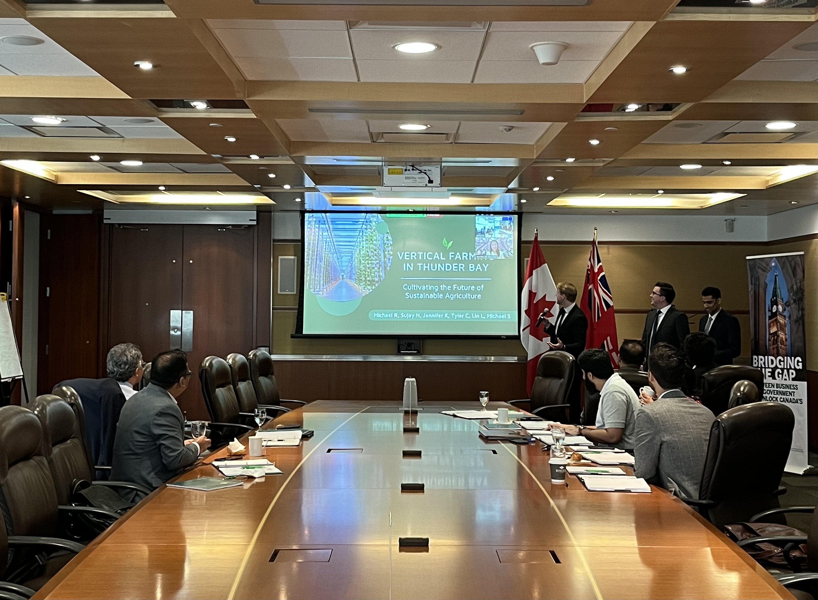 Long boardroom table with two judges on the left and five judges on the right. Three students stand at the front of the room to the right of a drop down screen. Screen is showing title page of power point deck and video feed of student participating virtually. All people in the room are looking at video feed.