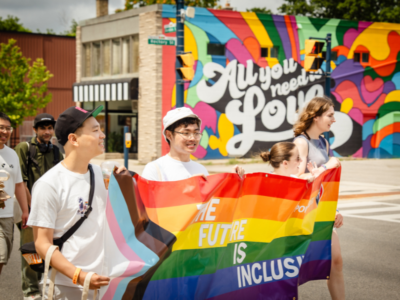 Ivey representatives walking with Pride flag that reads The Future is Inclusive. A rainbow mural with text All you need is love is in the background.