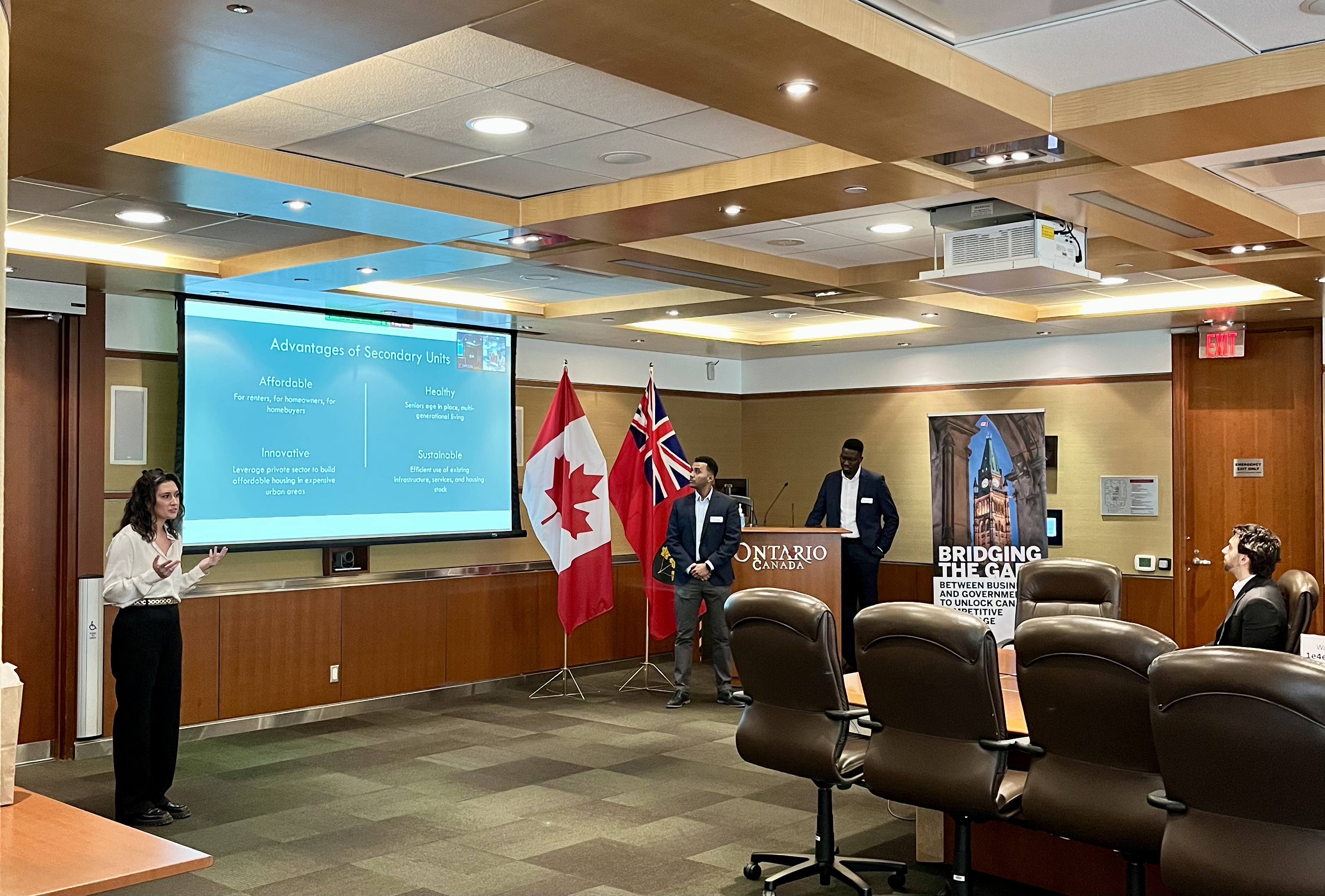 Three students standing at the front of a board room, one to the left of a drop down screen, the other two to the right. There is a podium with "Ontario Canada" written on the front.